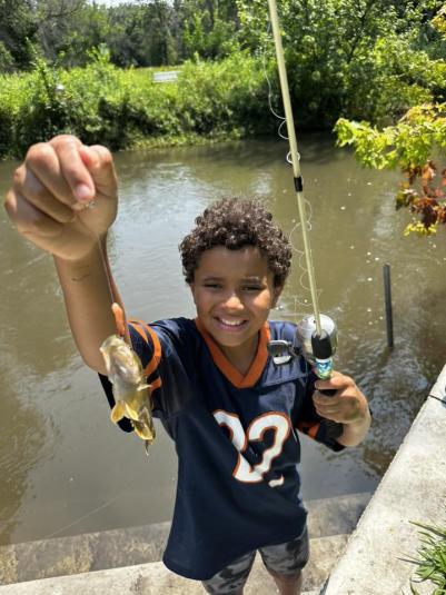 A boy stands in front of a body of water, holding up a fish he just caught.