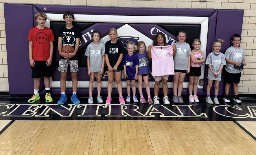 Eleven students stand in the gym, the backs against a hanging purple, black, and white mat that reads "South Central Calhoun." On the far left are the two older helpers.  Most of the younger players are either wearing or holding up T-shirts that read "SADA Confidence."