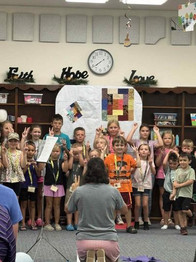 Approximately 18 children stand in front of a dark brown bookshelf, facing the camera. In front of them, with her back to the camera, is a woman kneeling on the floor, leading the children in song.