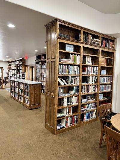 Part of the interior of the J.J. Hands Library in Lohrville, Iowa. The room is brightly light with white walls and ceiling, and brown flooring. In the foreground is a ceiling-to-floor wooden bookshelf that creates a half wall. In the background are more bookshelves.