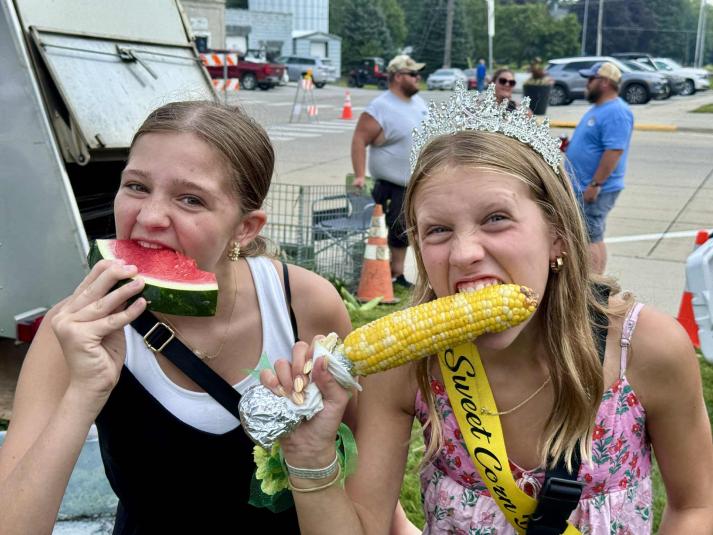 The girl on the left eats a slice of watermelon while the girl on the right eats sweet corn on the cob.