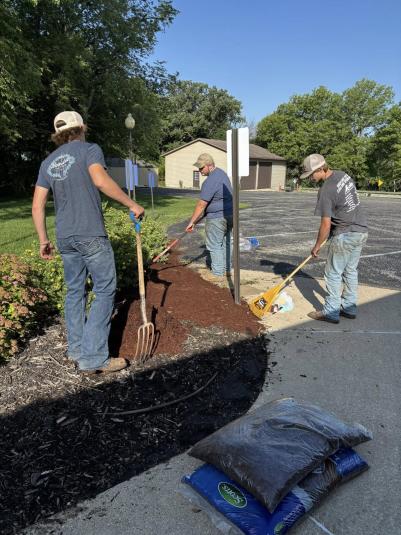 Three male South Central Calhoun FFA members work outside, raking mulch. They have their backs to the camera. Each is holding either a rake or pitchfork.