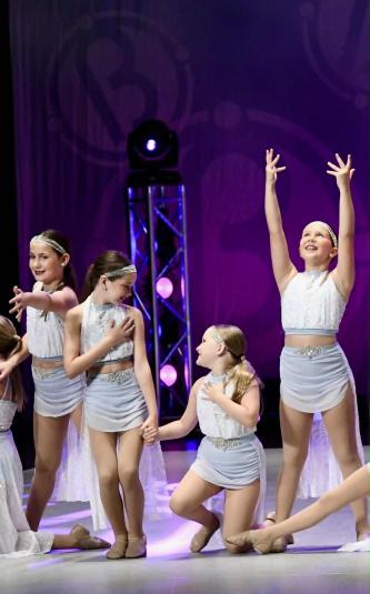 Four girls stand on stage wearing white sleeveless shirts and silvery grey skirts. The girl on the left is looking forward, reaching out with her left hand. The two girls in the center are holding hands, looking at each other. The fourth girl, on the far right, is looking up while reaching her hands to the sky.