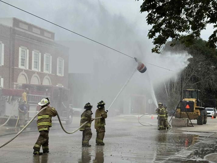 Two teams of firemen face each other, with an overheard marker between them. The teams are using their hoses to move the marker.