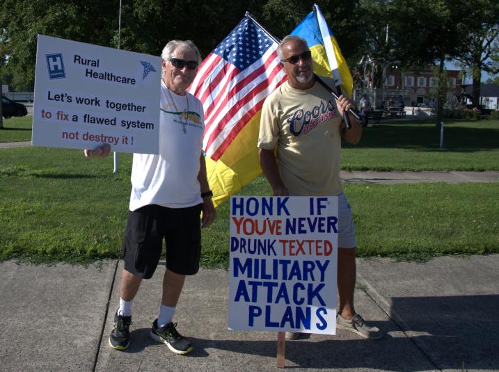 Two men stand facing the camera. The man on the left holds a sign saying "Rural Healthcare: Let's work together to fix a flawed system, not destroy!" The man on the right is holding a sign that says, "Honk if you've never drunk texted military attack plans." He is also holding an American flag and a Ukrainian flag in his left hand.