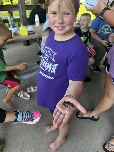 A girl stands facing the camera, holding a bug in her left hand.