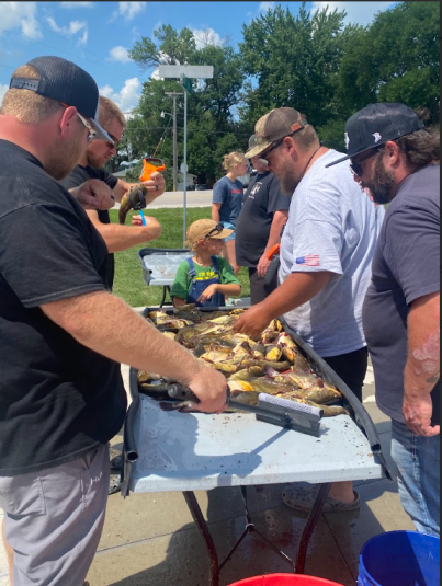 In the center is a white table with several caught bullheads. Standing on either side of the table, looking down at the fish, are two rows of men.