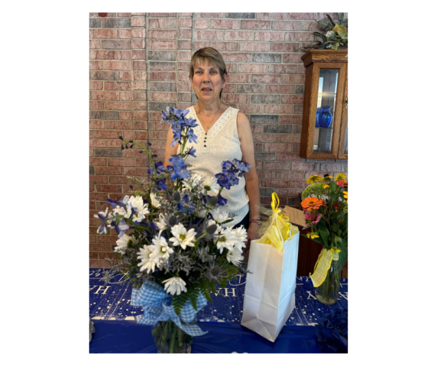 Mary stands in front of flowers and gifts set out on a table.