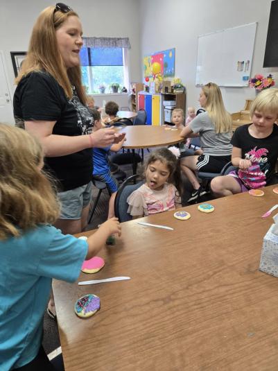 Gangelhoff stands in the library, next to three children sitting at a wooden table, who are frosting sugar cookies. In the background are two more tables with children who are also decorating cookies.