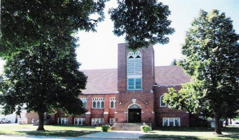 An outdoor photo of the red brick church taken on a sunny day. In the background is the church, with its three-story steeple. In the foreground are tree branches covered with leaves, which partially obstruct the view of the church.
