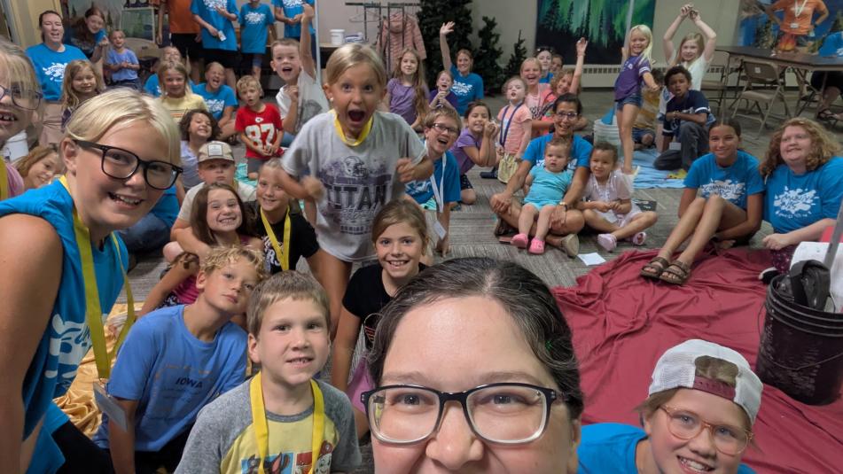 Brandt is taking a selfie and a room full of adults and children are behind her, smiling and waving at the camera.