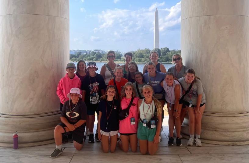 A group of students and chaperones stand outdoors, between two giant stone pillars. In the distance is the Washington monument.