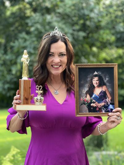 Debbie DeCamp stands outside, with green trees in the background. She is wearing a purple dress, holding a trophy in her right hand and her coronation photo in her left hand. She is wearing a tiara and smiling at the camera.