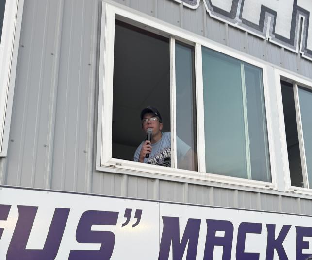 Greene sits in the press box in front of an open window, microphone in hand.