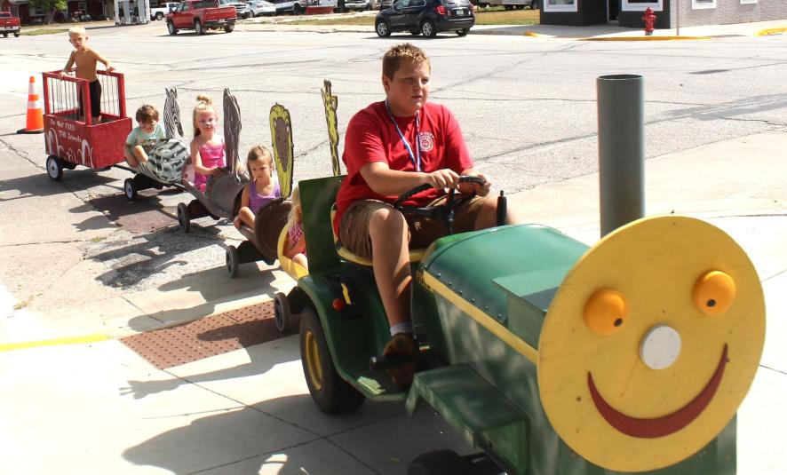A young man drives a lawn tractor while pulling several small cars with passengers.