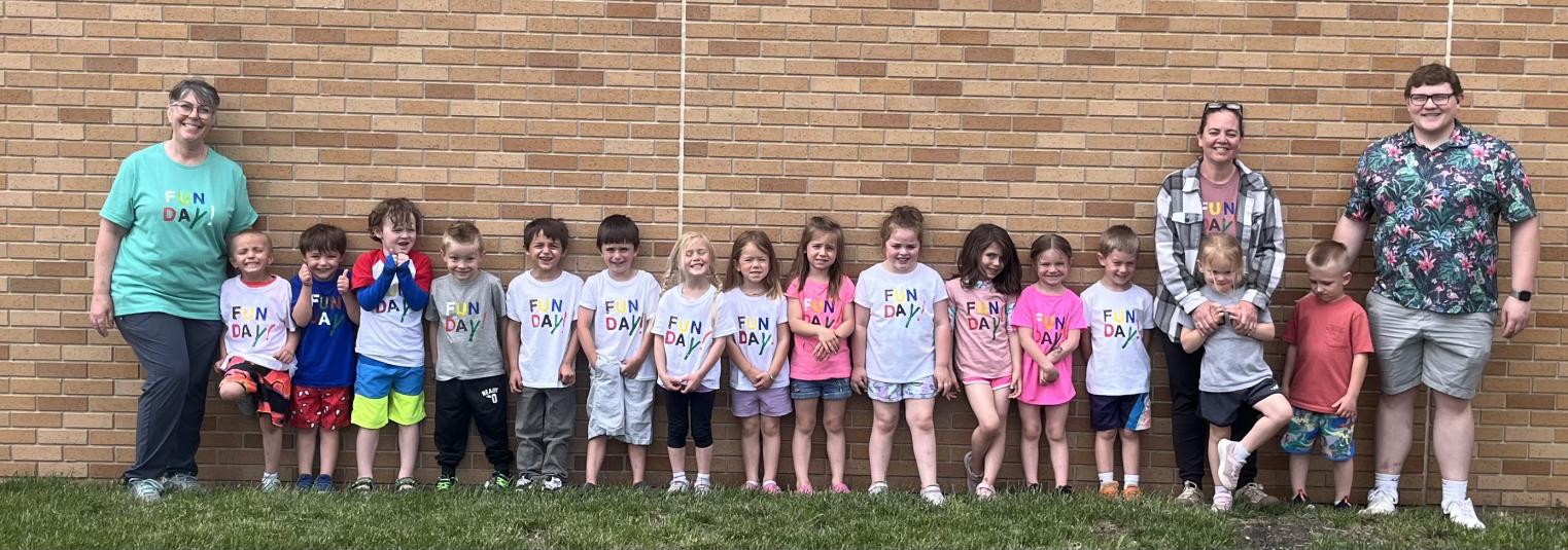Students and teachers stand outside against a brown brick wall. Most of them are wearing T-shirts that say "Fun Day" on the front.