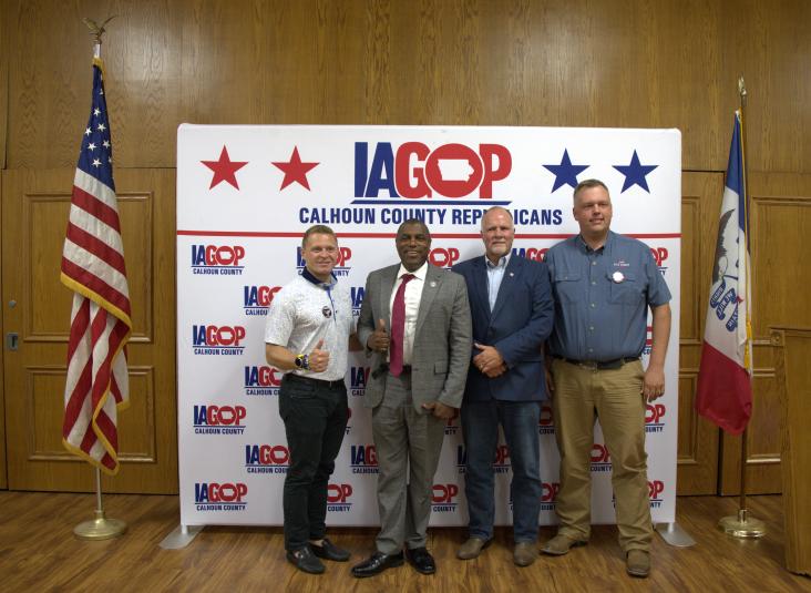 The four men stand against a photo opp background that reads "IA GOP." Bousselot is giving the thumbs-up sign.
