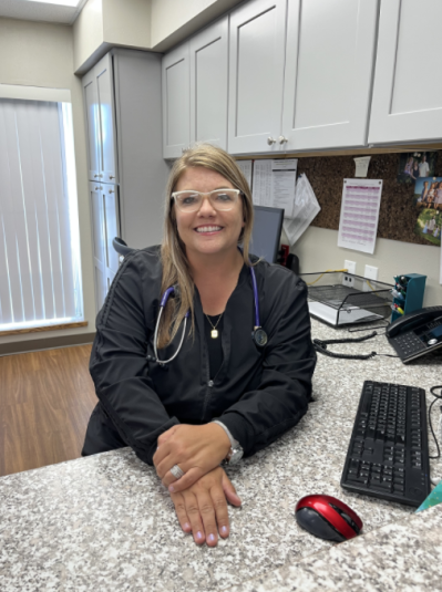 Holly sits behind her desk, smiling and looking at the camera. Her keyboard and mouse are visible. Cabinets are in the background.