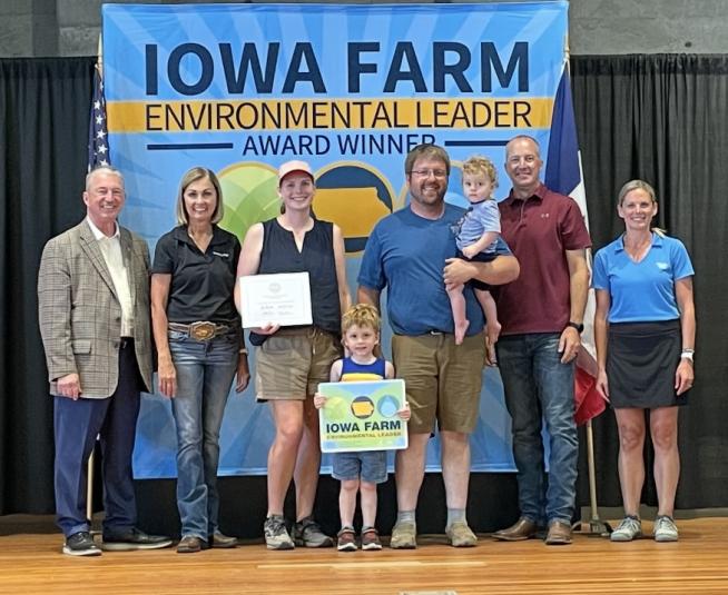 The Hepp Family stands on stage with Iowa Gov. Kim Reynolds, as well as three other unidentified individuals. Behind them is a backdrop that reads "Iowa Farm Environmental Leader Award Winner."