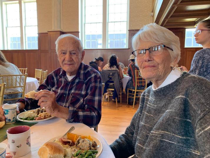 Jack and Carol are seated at a round table eating Thanksgiving dinner. They are both looking at the camera and smiling. 