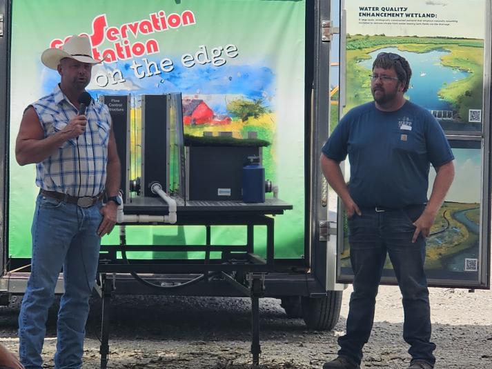 James Hepp stands in front of a large banner that says "Conservation Station: on the edge." To his left is a man speaking into a microphone.