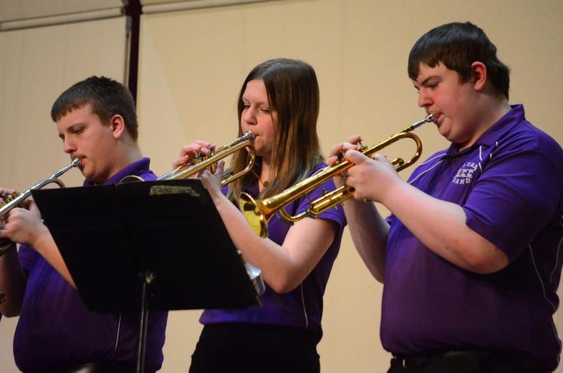 Three SCC students stand, playing the trumpet. From left to right are a boy, a girl, and a boy. All three are wearing purple polo shirts and black pants. They are looking at the music stand in front of them.