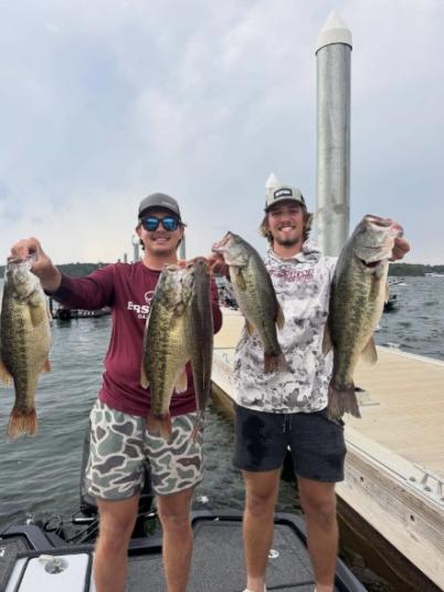 The two men stand next to a dock, both holding a fish in each hand.