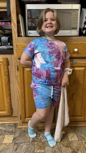 A girl stands in front of kitchen cupboards, wearing blue, white, and purple T-shirt and shorts pajamas.