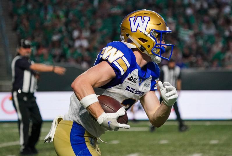 A close-up photo of Kody Case running, holding the football in his right hand. He is wearing a gold helmet with a white "W" on the side. The faceguard portion of the helmet is bright blue. His jersey is predominantly white with blue and gold accents. His pants are gold with a blue stripe down the side. In the background are two referees. 