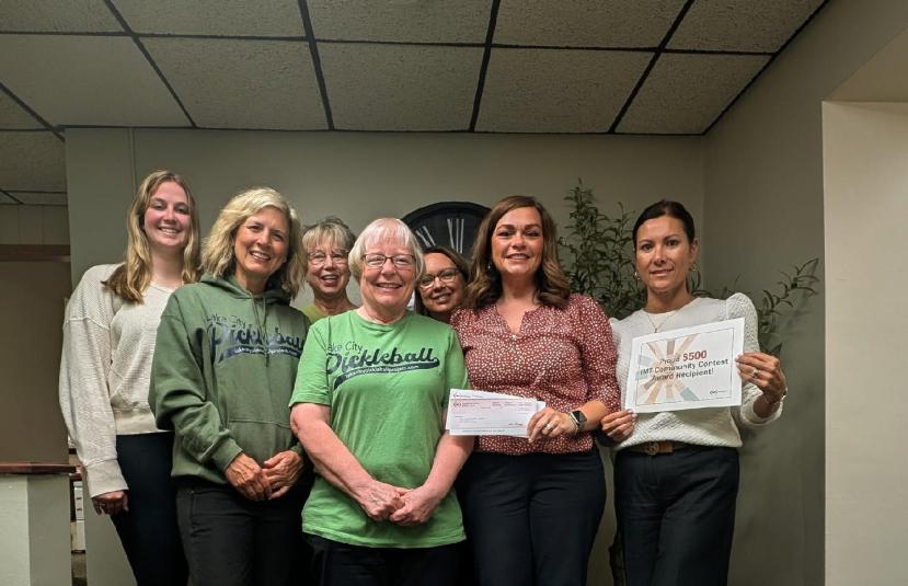 A row of seven women stands in two rows. The woman on the far right is holding a sign that says "Proud $500 IMT Community Contest Award Recipient!" The woman to her right is holding a check.