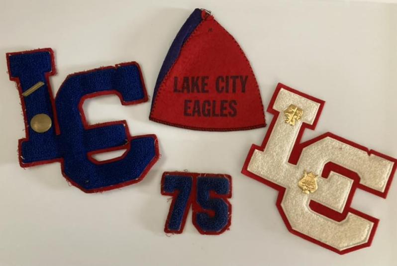 Lettermen's jacket patches from the Lake City, Iowa, HS Class of 1975. Starting clockwise from the top, a red felt triangle that says "Lake City Eagles" in navy blue print; a white "LC" set against a red trim background; a navy blue "75" set on red trim, and a navy blue "LC" set on a red trim.
