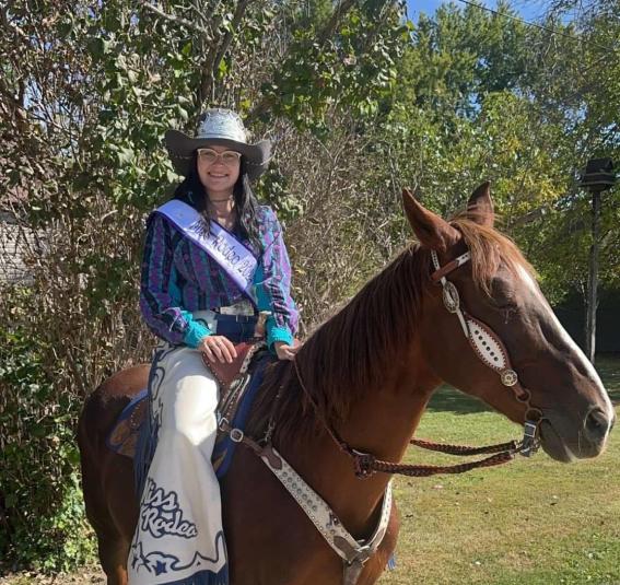 Lily Genter is outdoors on a sunny day, sitting on a brown horse. She is looking at the camera. In the background are green trees.