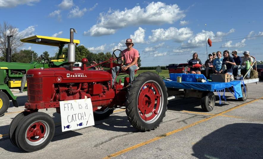 Buse sits on a red tractor that is pulling a flat-bed trailer with eight FFA members seated on it. A sign "FFA is a Catch!" is hanging on the side of the tractor. Several of the FFA members are holding fishing poles and fake fish.