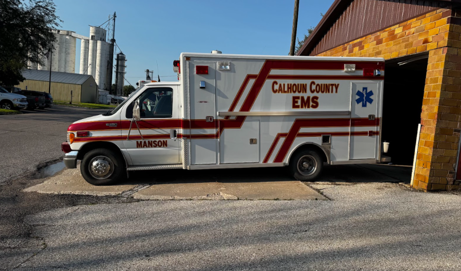  A side view of a white ambulance with red and gold stripes. The word "Manson" is on the driver's door, and "Calhoun County EMS" is on the rear side.