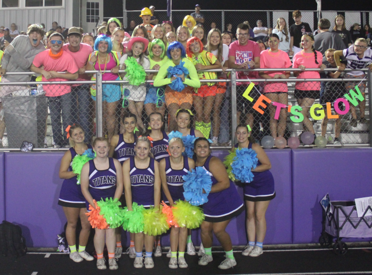 Cheerleaders stand on the ground in front of the student section of the bleachers. Many students are wearing neon colored clothing.