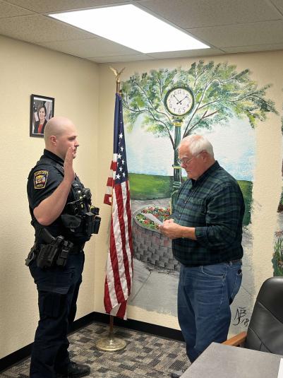 A side view of Lake City Chief of Police Jason Gray and Mayor Mike Schleisman standing, facing each other. Gray is in uniform and raising his right hand. The mayor is looking down at a piece of paper he is holding.
