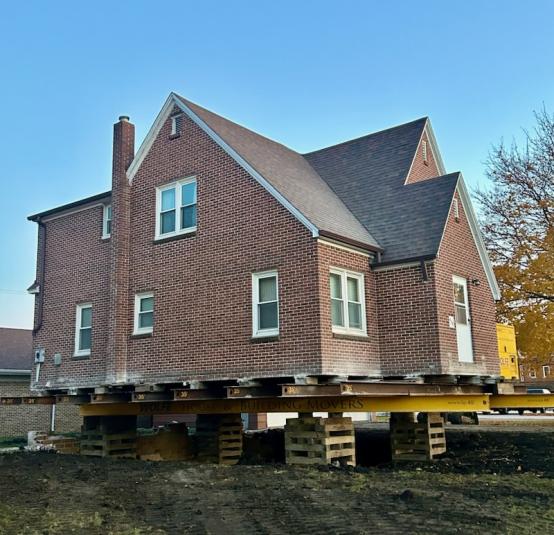 The two story, brownish red brick building is lifted off its foundation. Several wooden pallets are supporting it.