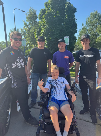 Rylan sits in a wheelchair, giving the thumbs-up sign with his right hand. Behind him are four men.
