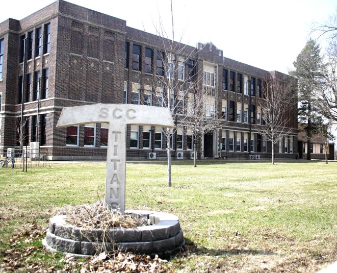 A far away shot of the exterior of South Central Calhoun High School in Lake City, Iowa, taken on a sunny summer day. The sun is shining and the grass is green. In the background is the three-story brown brick building. In the foreground is a metal "T" shaped lawn decoration that says "Titans."