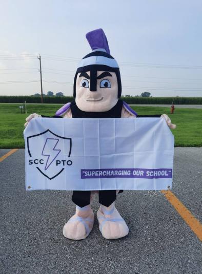 An individual wearing the Titan mascot costume is standing in a parking lot, holding a white flag that reads "SCC PTO: Supercharging our school"