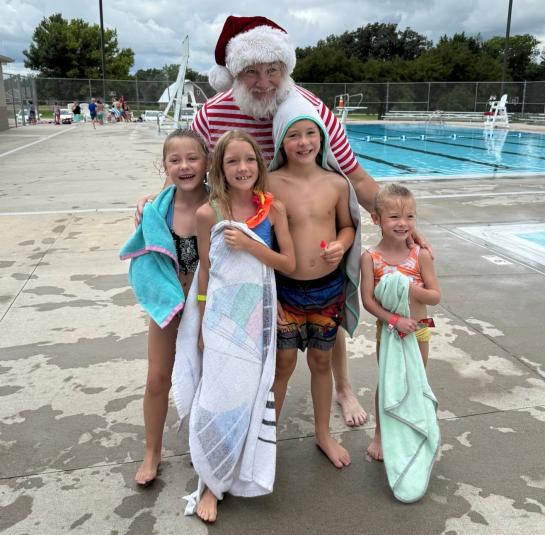 Santa Claus, wearing a red and white striped T-shirt and shorts and classic Santa hat, stands behind three children wearing bathing suits and holding towels. One boy is also wearing a Santa hat. In the background is the pool.