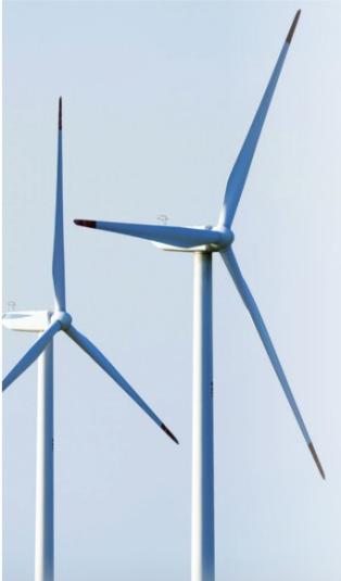 A photo of two grey wind turbines, close-up to the blades. The sky is clear and blue.