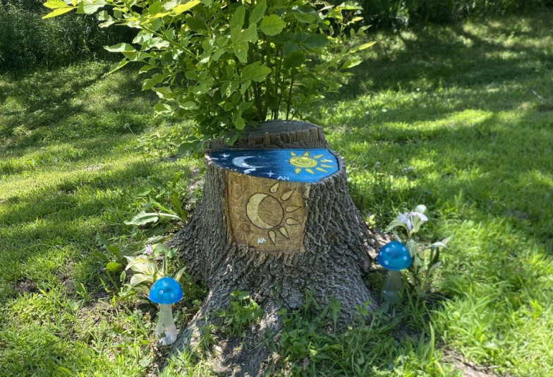 A photo of the decorated tree stump, taken on a sunny day. In the foreground is the stump. A portion of the bark has been peeled away, revealing a carving of an eye. On the top of the stump is a painting of the sun and moon set against a blue sky. Next to the stump, on either side, are two whimsical blue and white mushrooms, either made of plastic or glass. In the background is green foilage.