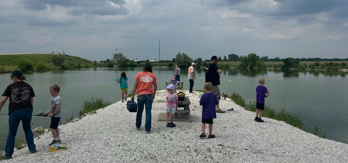 Approximately 10 adults and children stand on a gravel peninsula that juts into the lake. Several of them are holding fishing poles.