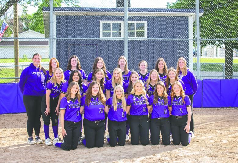 The South Central Calhoun softball team poses outside on the field, with their coaches. They are in three rows, with the first row kneeling and the back two rows standing. They are wearing purple jerseys and black pants.