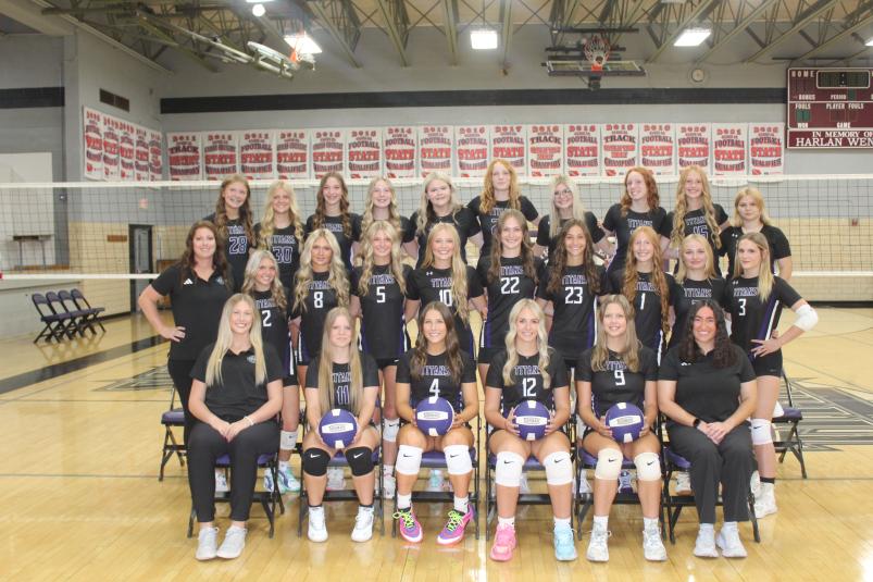 The players in uniform pose in the gym in front of the volleyball net, along with their coaches.