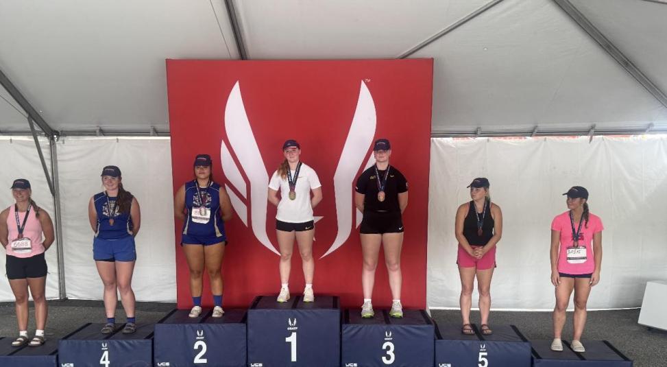 The top seven athletes stand on raised navy blue podiums. Each podium is numbered one through seven. Number one is in the center. Six, four, and two are on the left. Three, five, and seven are on the right. Each athlete is wearing a medal around her neck.