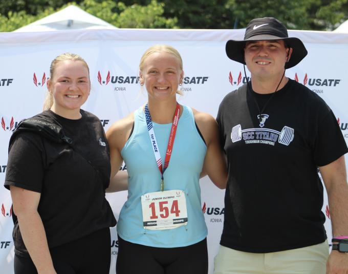 Tristin Gorden stands in front of a white "photo op" wall that has a repeating pattern of "USATF Iowa" on it.  She is wearing a blue tank top, black pants, and a lanyard that says, "1st place." Hanging off the lanyard is a sign that reads "Junior Olympic 154." She is standing between a man and a woman. All 3 are looking at the camera and smiling.