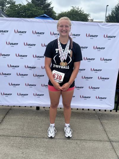 Tristin Gorden stands outdoors, facing the camera. She has on a black T-shirt, pink shorts, and has 3 medals around her neck. 