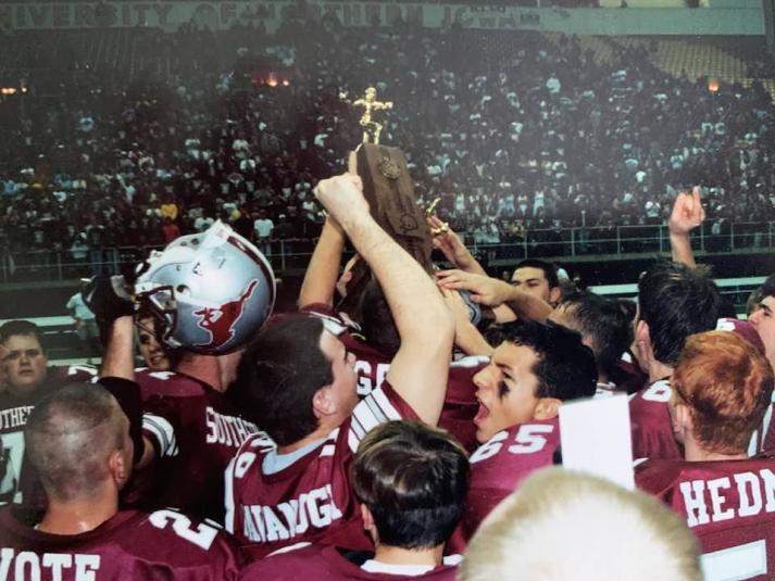 Several players are huddled close together, wearing maroon jerseys that say "Mustangs." A player in the center of the group is holding the trophy above everyone's heads. 